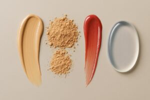 A flat lay of four makeup textures arranged on a beige background: a creamy foundation smear, two mounds of loose powder, a streak of glossy red lip gloss, and a smooth silicone sponge, highlighting different tactile experiences for sensory-sensitive users.